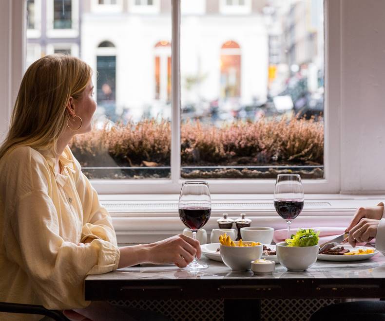 A window-side table for two at Jansz. with wine glasses and a woman enjoying the view of the streets of Amsterdam.