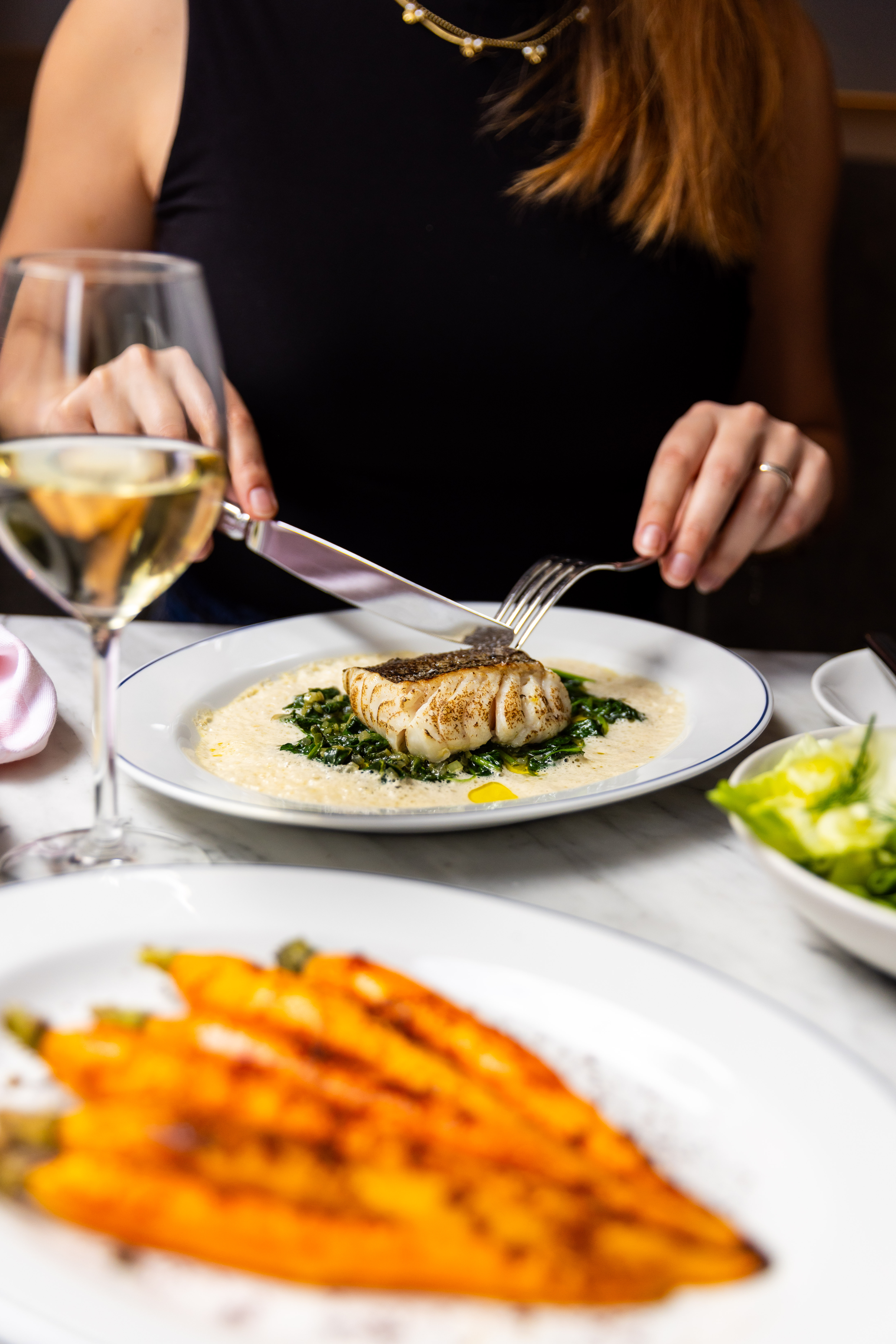 A woman cutting into a grilled fish fillet with vegetables on a plate, with a glass of white wine on the table at Jansz.